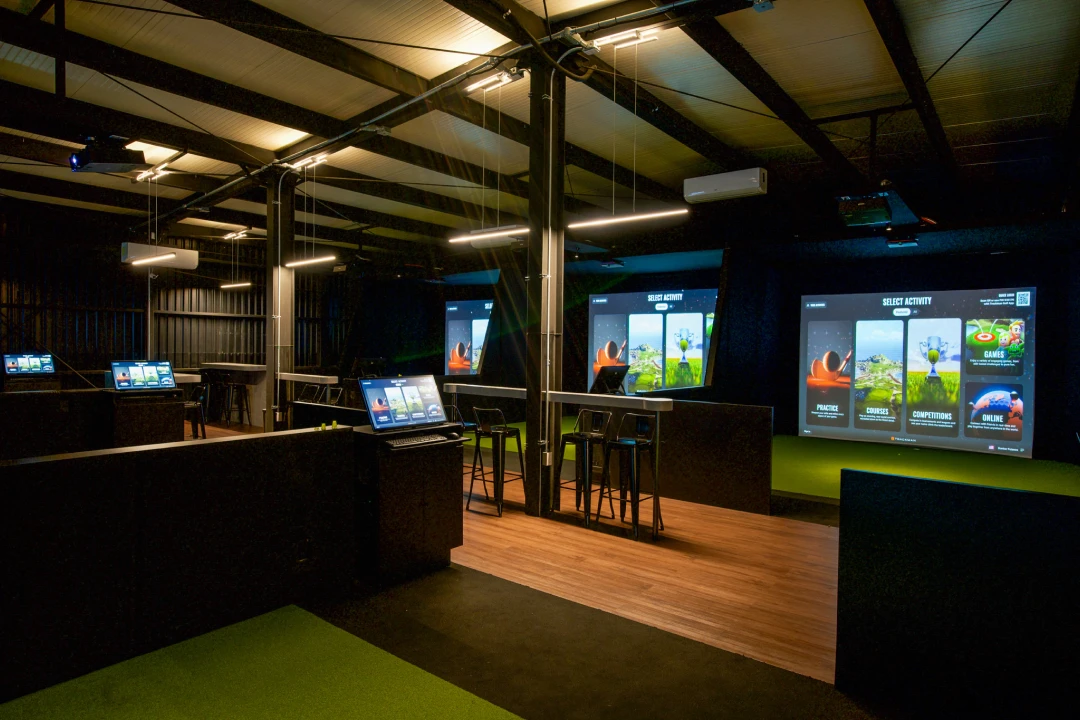 A dimly lit indoor room with multiple high-tech golf simulators, chairs, and screens displaying various activity options.