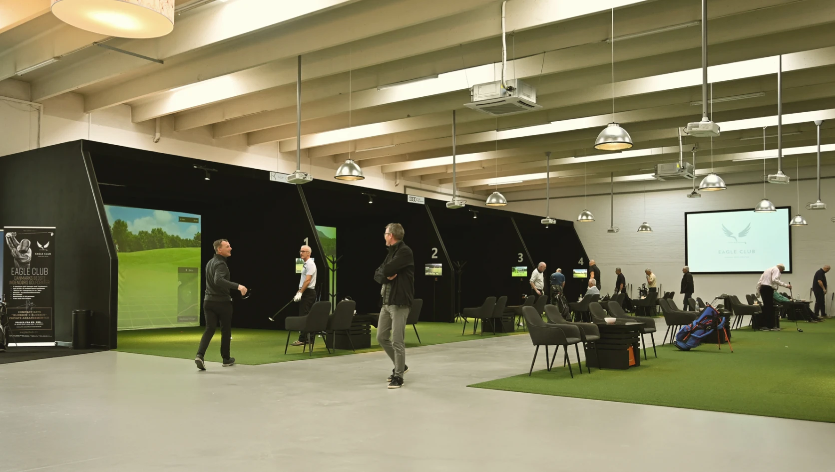 Indoor golf facility with people practicing at multiple simulator bays. Black dividers, green turf, and chairs are visible under modern lights.
