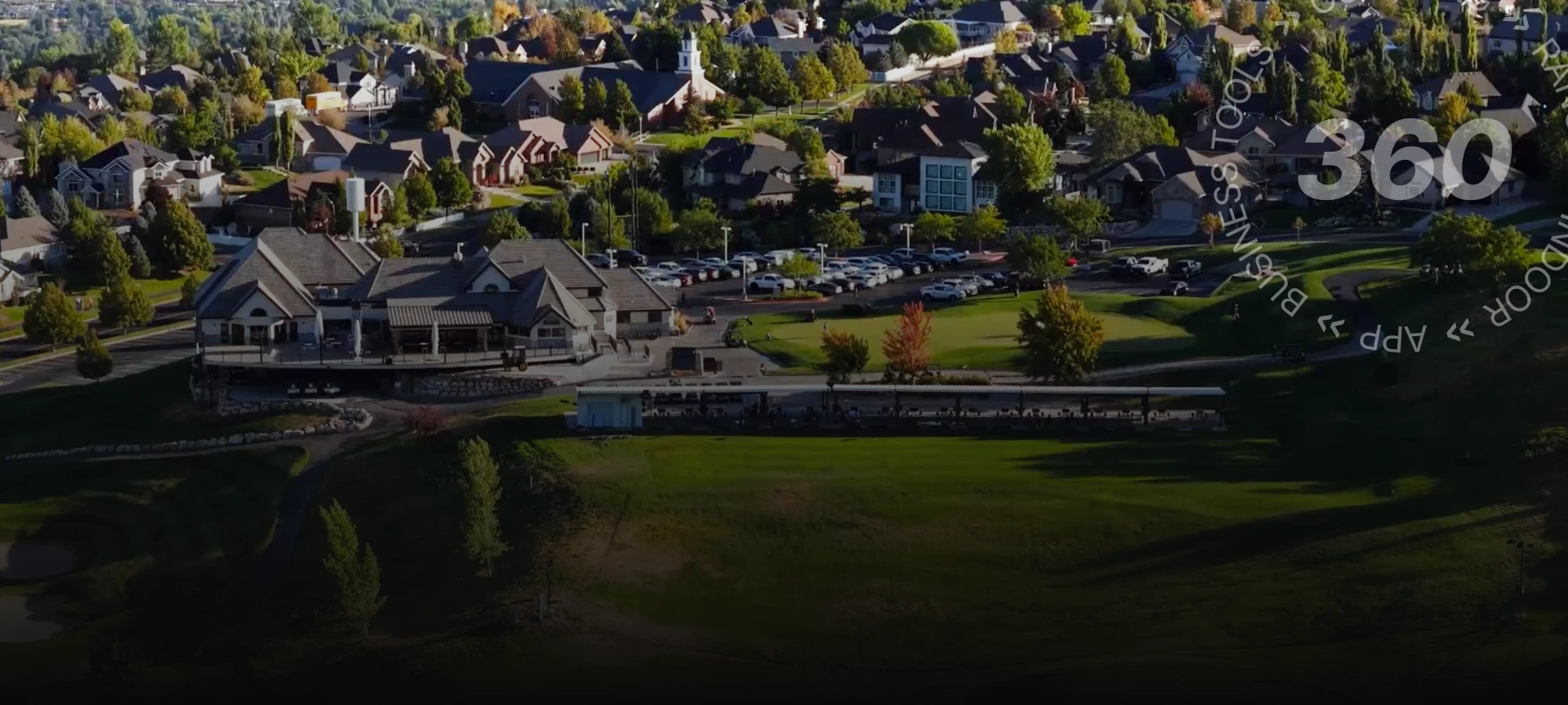 Aerial view of a suburban neighborhood with houses, a parking lot, and green spaces, surrounded by trees.