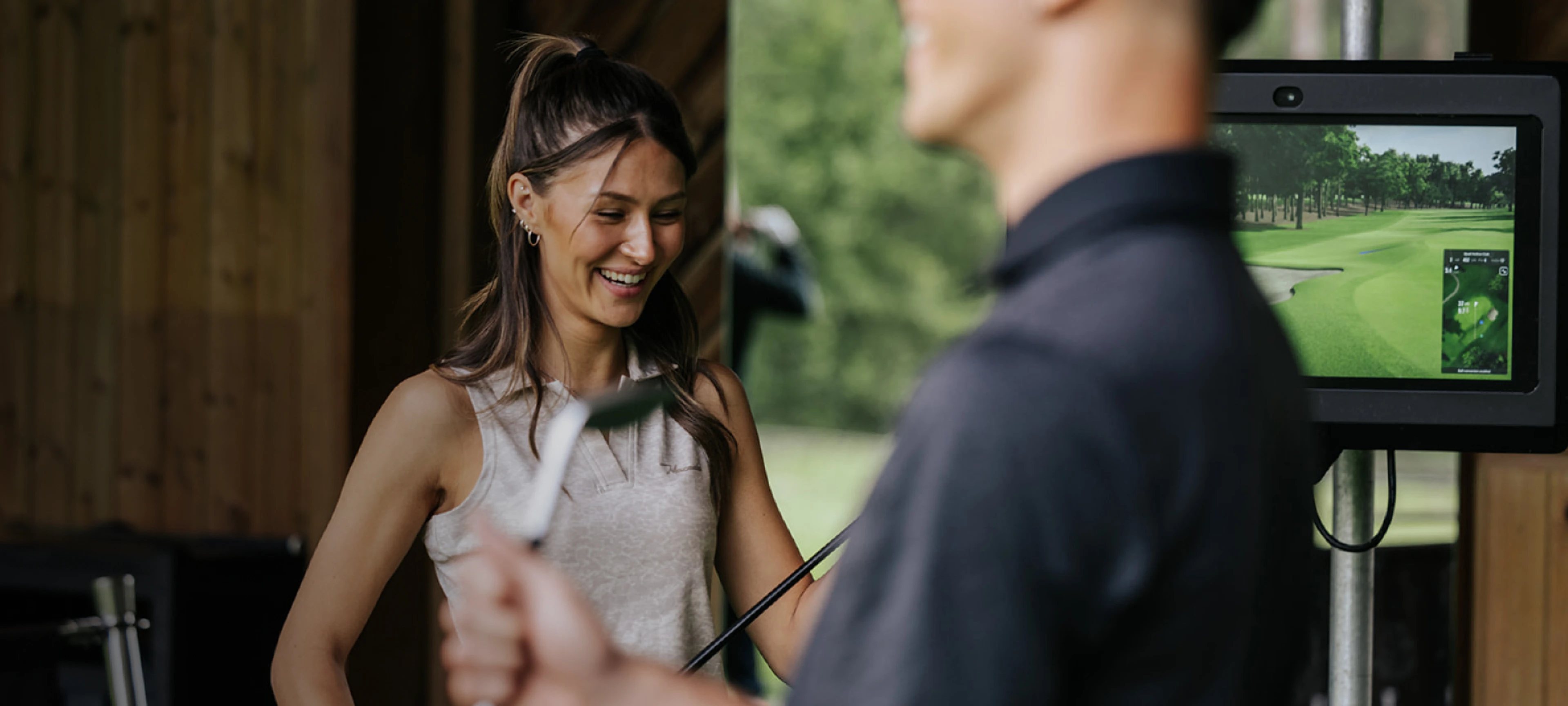 A woman smiling while holding a golf club indoors, with a man in the foreground and a virtual golf course on a screen in the background.