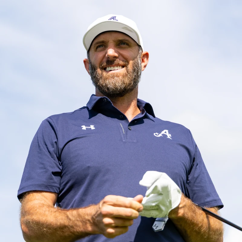 Smiling man in a navy golf shirt and cap holding a glove, standing outdoors against a blue sky.