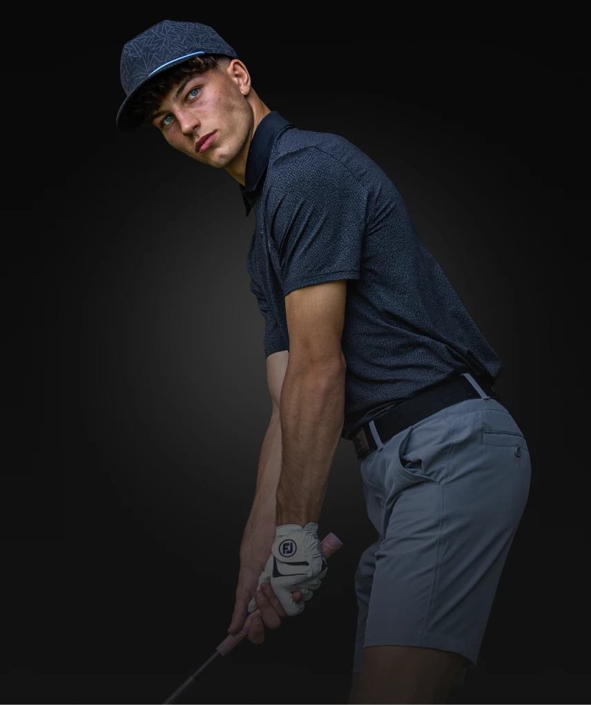 A golfer in a dark shirt and gray shorts, holding a golf club and wearing a cap, poses against a dark background.
