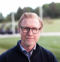 A person with glasses, light beard, and short hair stands outdoors, wearing a dark jacket with a grassy field and trees in the background.