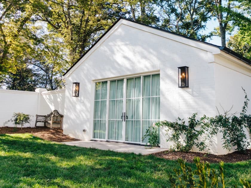 White building with large glass doors, flanked by wall lanterns, surrounded by greenery and trees, and a wooden bench nearby.