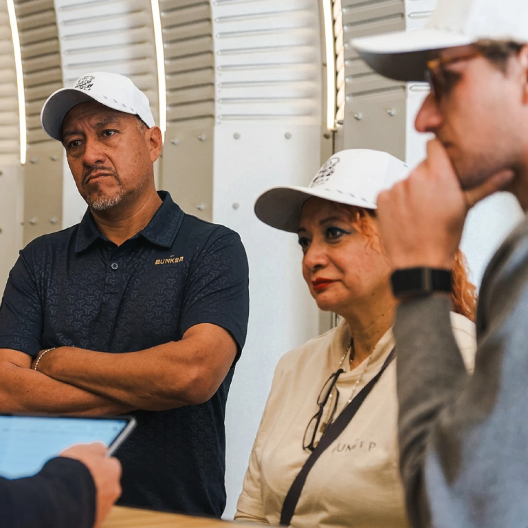 Three people in casual attire and caps attentively listen to someone off-camera, inside a modern, well-lit room. One appears to take notes.