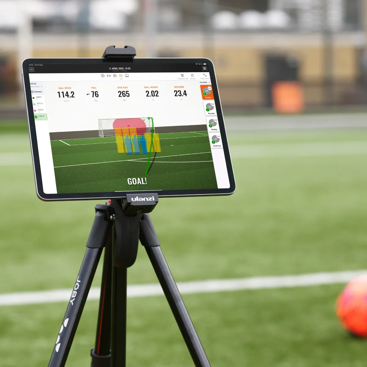 Tablet on a tripod displaying soccer goal statistics on a field, with a blurred orange ball nearby.