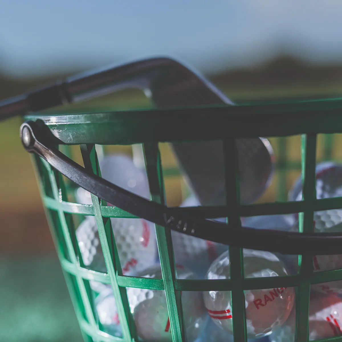 Basket of golf balls with an iron club on a grassy field, under a clear blue sky.