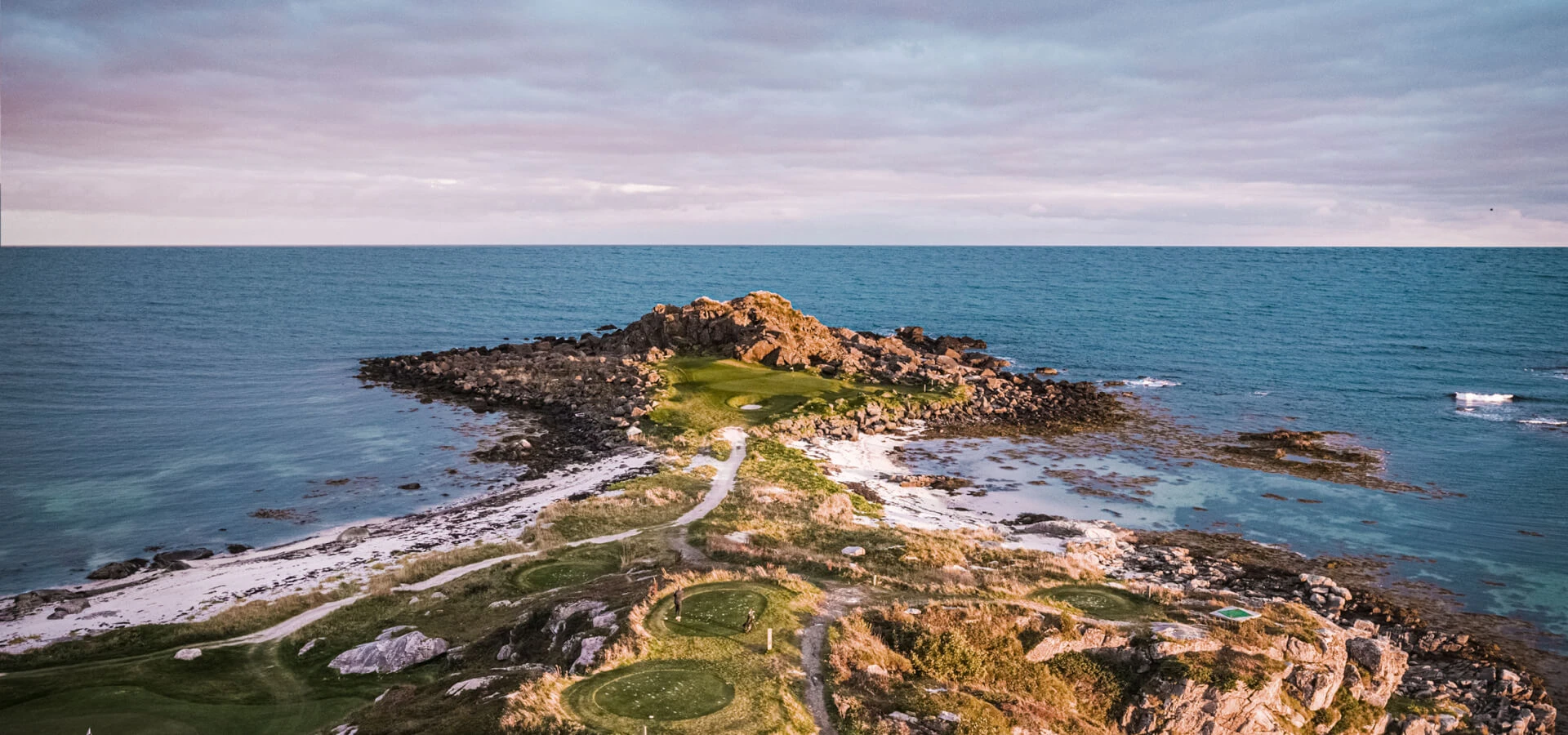 Aerial view of a rocky coastline with a narrow path leading to the sea, surrounded by calm ocean waters under a cloudy sky.
