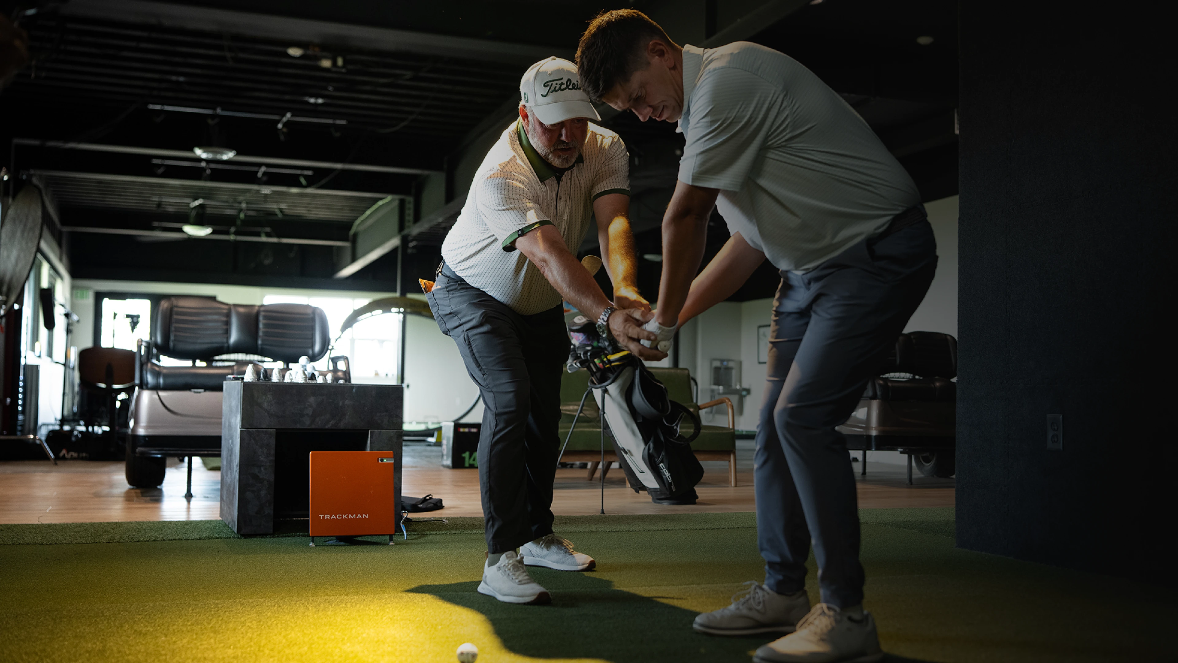 Golf instructor guiding a student on swing technique indoors, with equipment and seating visible in the background.
