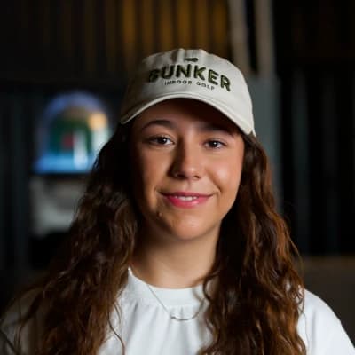 A woman with long brown hair wearing a "Bunker Indoor Golf" cap and white shirt, smiles softly against a dimly lit background.