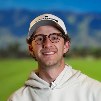 A smiling person in glasses and a cap stands outdoors with a blurred scenic background of hills and a bright blue sky.