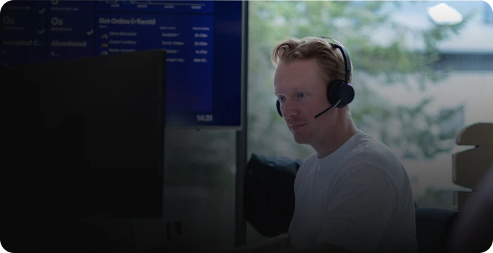 Person wearing a headset, sitting at a desk with a computer screen, faint data displayed in the background.