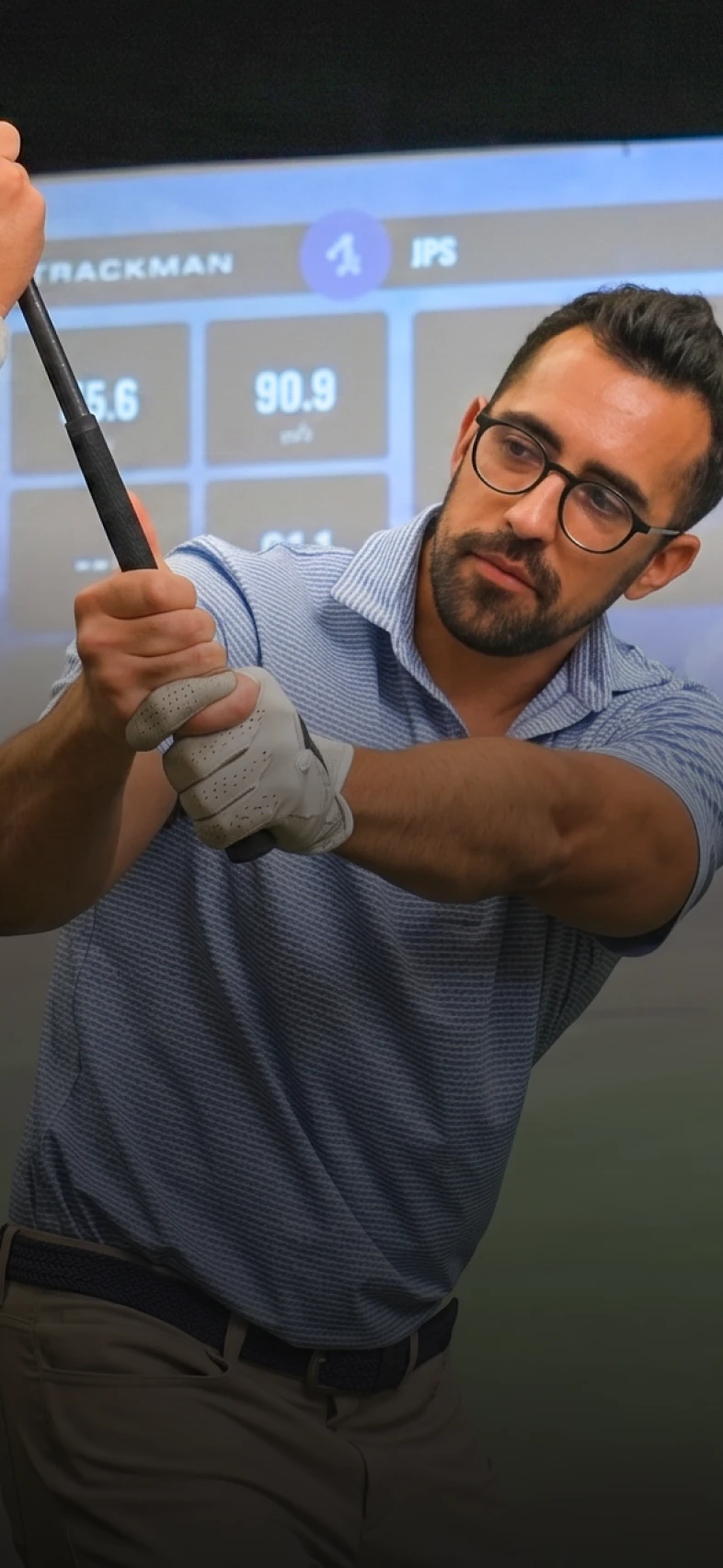 Man in glasses gripping a golf club indoors, wearing a light blue striped shirt and gloves, with a screen displaying data in the background.