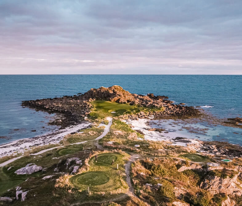 Aerial view of a rocky coastal landscape with a narrow path leading to a grassy area, surrounded by the ocean under a cloudy sky.