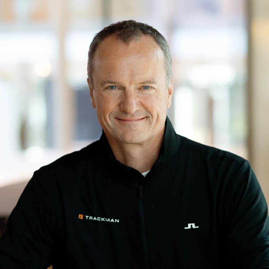 Smiling man in a black jacket with logos, standing indoors with a blurred background.